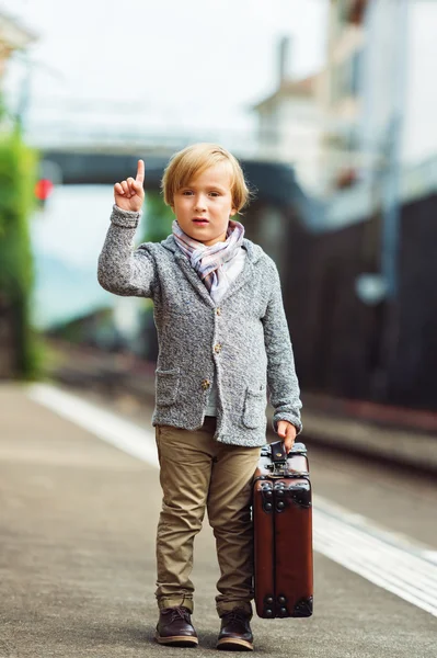 Adorable little boy on a railway station, waiting for the train with suitcase, checking wind direction with his finger