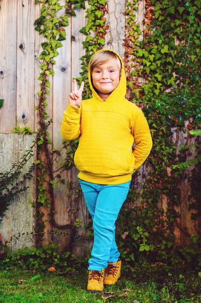 Outdoor portrait of adorable little 5 year old blond boy playing in the park, wearing yellow sweatshirt with hood, shoes and blue trousers