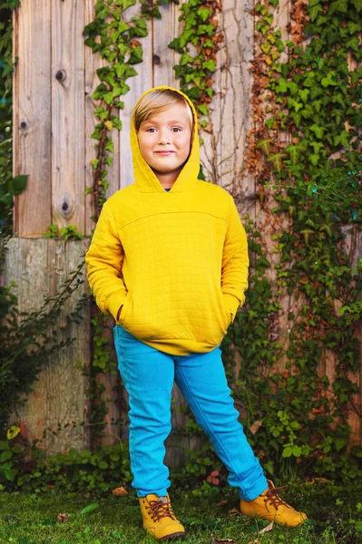 Outdoor portrait of adorable little 5 year old blond boy playing in the park, wearing yellow sweatshirt with hood, shoes and blue trousers