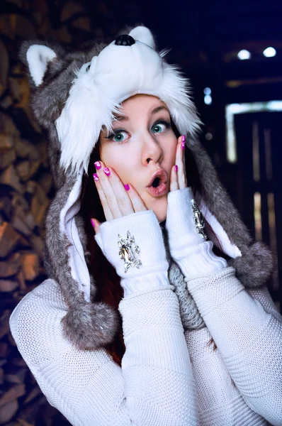 Teenage girl with red hair and green eyes wearing a furry husky hat with ears and pompoms, and knitted hand warmers with diamonte detail is touching her cold cheeks with a surprised expression on her face.