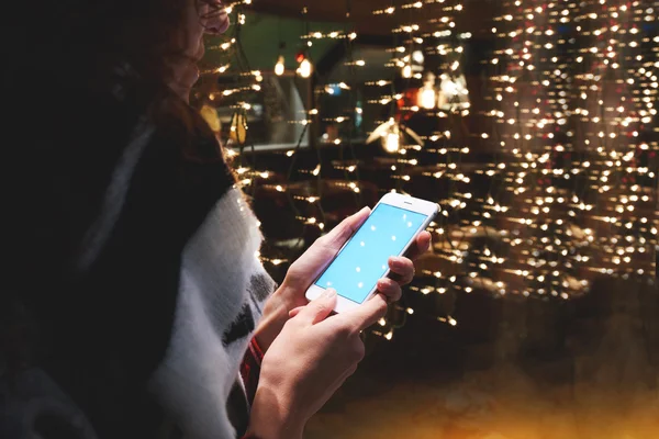 Closely of woman is reading text message in social network via smart phone, while is standing outdoors against garlands lights.