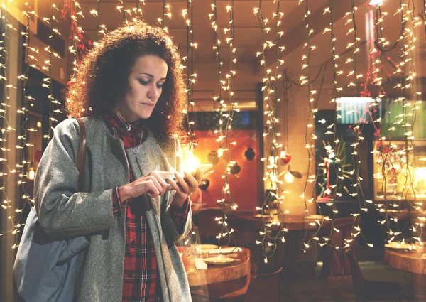 Young woman is reading text message on cell telephone from her boyfriend, while is standing against garlands lights in Valentine\'s day.