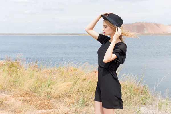 Beautiful young blond woman in a black dress and a light black hat in the desert and the wind blowing her hair in a hot summer day