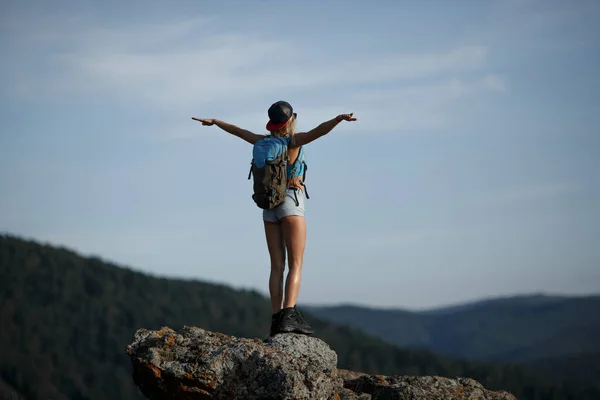 Hiking happy woman with backpack standing on rock with raised ha