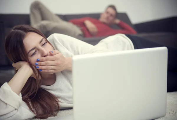 Woman yawning at laptop and man lying on couch