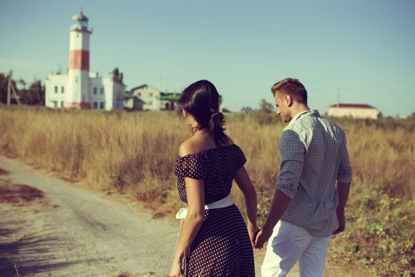 Young couple hipster indie style in love walking in countryside, holding hands, lighthouse on background, warm summer day, sunny, bohemian outfit, vintage bag with flowers. Copy space