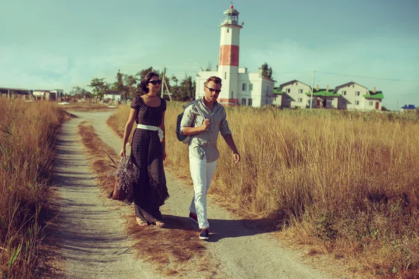 Young couple hipster indie style in love walking in countryside, lighthouse on background, warm summer day, sunny, bohemian outfit, vintage bag with flowers