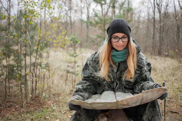 Young beautiful woman in camouflage outfit sitting on forest log discovering nature in the forest with compass and map. Travel lifestyle concept.