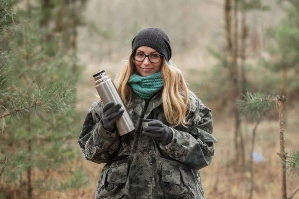 Young beautiful blond woman in camouflage outfit and green scarf posing with thermos in the forest. Travel lifestyle concept.