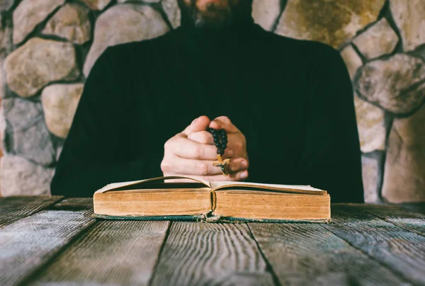 A man in black clothes with a prayer beads in hand praying in front of an old open book. the concept of praying, and studying. selective focus