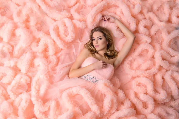 Beautiful young girl with long wavy hair lies on the pink dress-the cloud looks at me, view from above