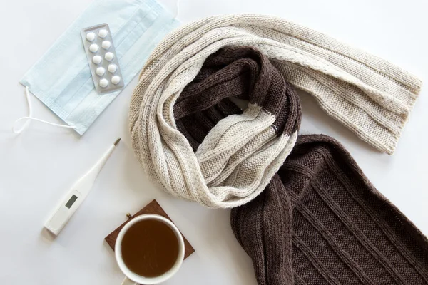 White desk with pills, thermometer, hot drink and a scarf on it