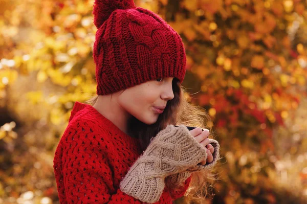 Happy child girl in warm red knitted hat and scarf on cozy autumn walk drinking hot tea from thermos. Seasonal outdoor activities