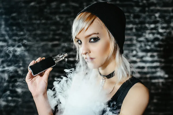 Sexy goth girl smokes electronic cigarette on dark background. The model vaper vaping a vaporizer in the studio.