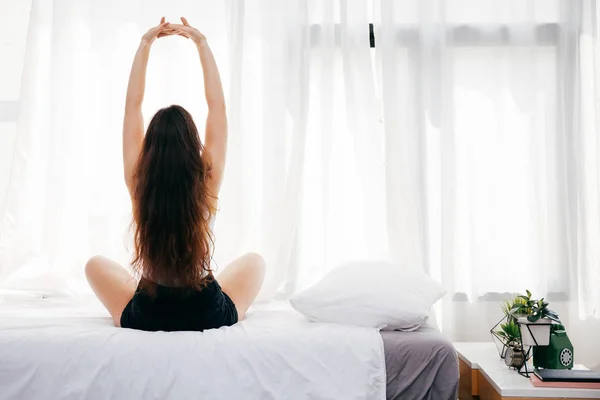 Back of young woman sitting and stretching up on bed in the morning