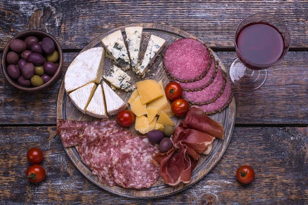 Red wine in a glass and a set of products - cheese, sausage, salami, olives, tomatoes, hot peppers, bread on a wooden board, background. Top view