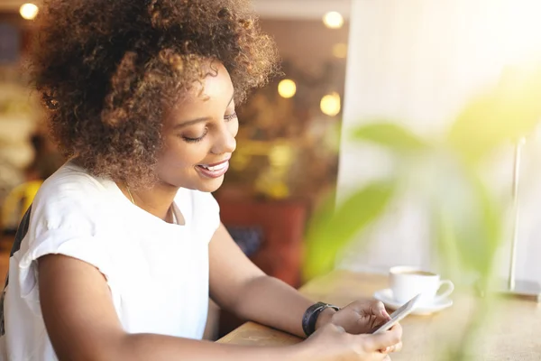 Fashionable African student girl with stylish curly hair using high-speed Internet connection, checking news feed, liking posts with happy smile, having cappuccino, relaxing at cafe after college