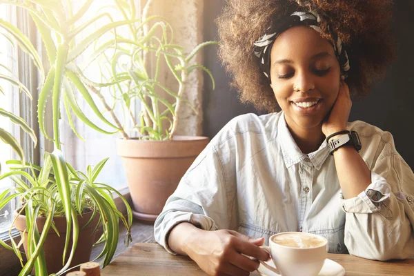 Young, beautiful and tender African female hipster sitting at home table near windowsill with two big flowerpots, holding mug with cappuccino, touching her neck with hand, looking down with shy smile