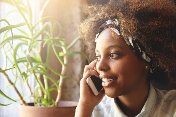 Close up portrait of attractive stylish dark-skinned girl with Afro hairstyle wearing bandana talking on mobile phone to her boyfriend looking aside, isolated against home interior background
