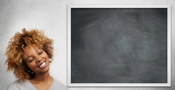 Back to school. Portrait of beautiful stylish student girl with Afro haircut, standing at blackboard, smiling and closing her eyes with happy pleased expression, dreaming about summer vacations
