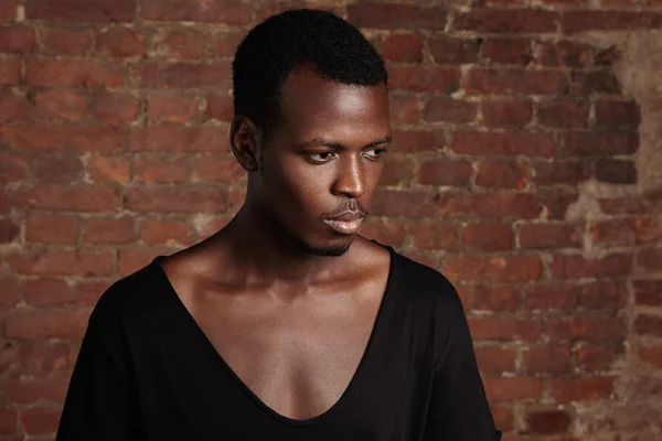 Close up studio shot of fashionable attractive pensive African male wearing stylish black t-shirt with low oval-shaped neck looking aside with serious and thoughtful face expression. Horizontal