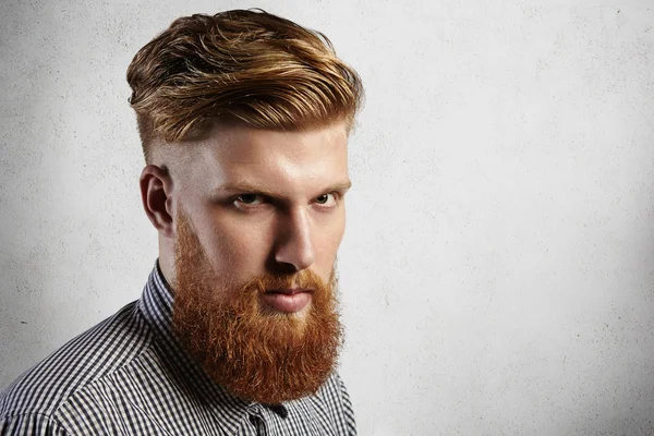 Manful European hipster in checked shirt looking seriously at the camera. His stylish haircut and well-trimmed blond beard say that he is loyal client of barbershop and takes care of his appearance.
