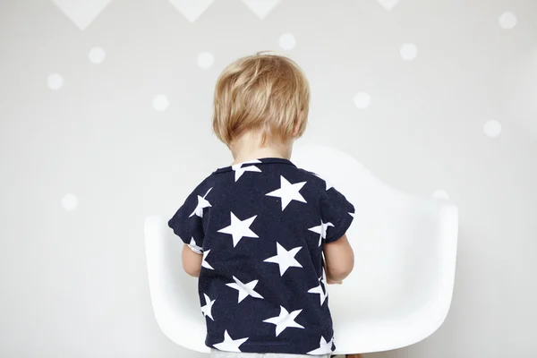 Back view of little Caucasian boy with blonde hair wearing t-shirt with stars on it, playing with toys in nursery. Cute toddler standing in front of white chair, spending day at home. Visual effects