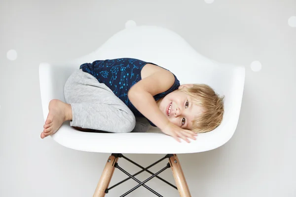 Childhood and leisure concept. Sweet adorable two-year old toddler with cute smile curling up in white chair, hiding himself while playing hide-and-seek with friends at nursery or kindergarten