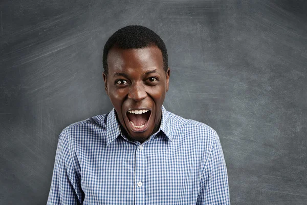 Negative human face expressions and emotions. Mad and furious young dark-skinned teacher standing at blank chalkboard, yelling at his disobedient students, calling for silence, screaming: Shut up!