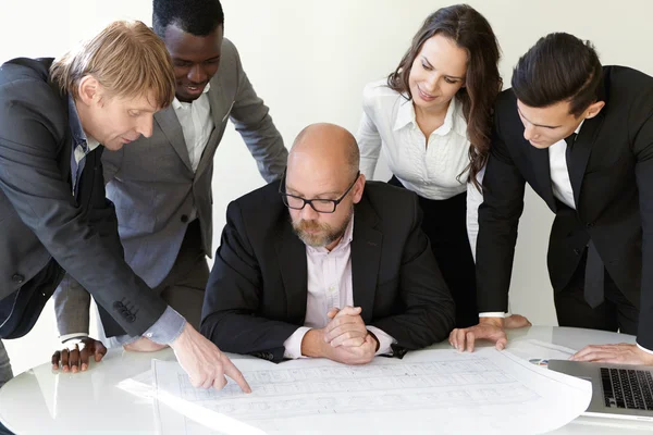 Team of architects studying blueprints during meeting while working on new engineering project. Young constructor pointing finger at drawings on table, showing sketch to colleagues and boss in glasses