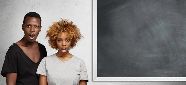 African man and woman standing close to each other at blank chalkboard with copy space for your text or promotional content, looking surprised and shocked, opening mouths widely in full disbelief