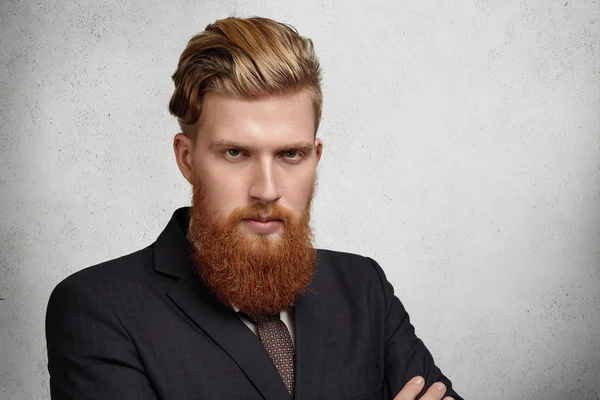 Headshot of good-looking young corporate worker with trendy haircut and long red beard dressed in elegant suit looking with focused and concentrated face expression, ready for serious business meeting