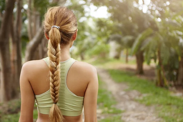 People, sports, nature and fitness. Close up rear view of young blonde woman runner with long braid dressed in sportswear standing on trail in green forest, ready to run while preparing for marathon