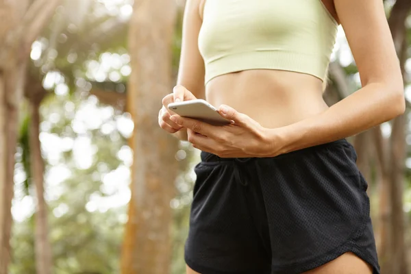 People and technology concept. Cropped shot of beautiful athlete woman with fit healthy body browsing newsfeed on social networks using Internet on her cell phone, standing against green trees