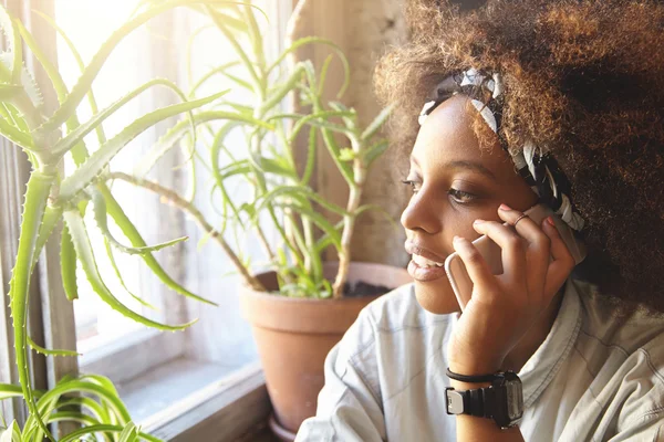 People, technology and communication concept. Cute fashionable dark-skinned woman with Afro hairctyle wearing white t-shirt, having phone talk with her friend and making appointment using mobile