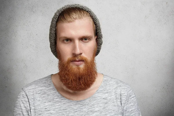 People, style and fashion concept. Isolated portrait of young handsome hipster model with stylish beard and mustache wearing gray warm knitte hat looking at camera with severe or angry expression