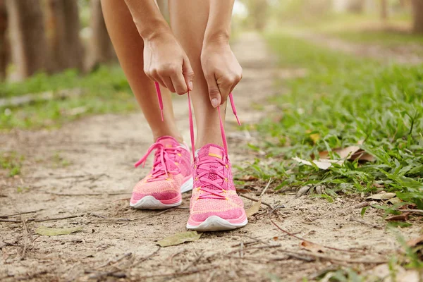 Cropped shot of young woman runner tightening running shoe laces, getting ready for jogging exercise outdoors. Female jogger lacing her pink sneakers standing on forest path before morning run