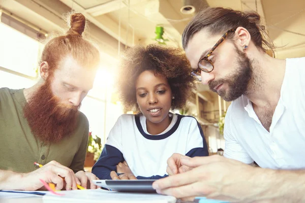 Creative people brainstorming at meeting using touch pad to note ideas and plans. Man in glasses showing presentation to African woman on tablet while their redhead colleague writing something down