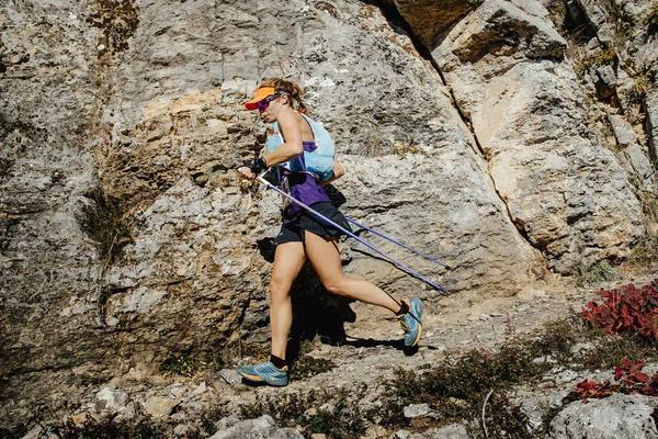 Woman runner with nordic walking sticks running trail on background of rocks
