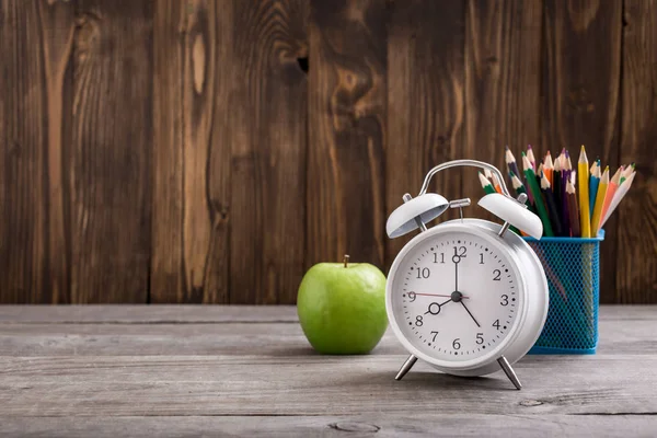 Alarm-clock with colored pencils and green apple on wooden table