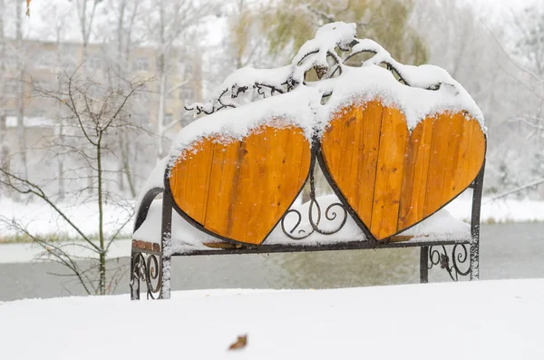 Empty snow-covered wrought-iron bench in the form of hearts for couple of lovers in winter park.