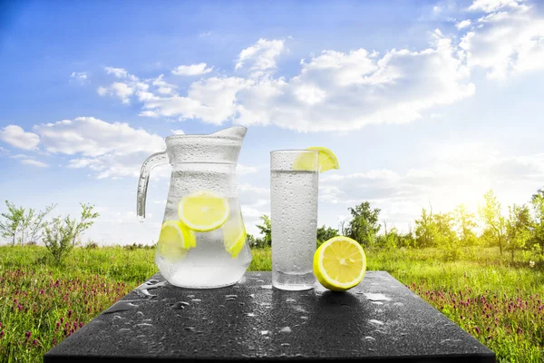 Fresh cold water with lemon and ice in a pitcher on the table.Homemade lemonade with fresh citruses on the background of nature. Quenching thirst.The condensation on the jug and a glass of cold water.