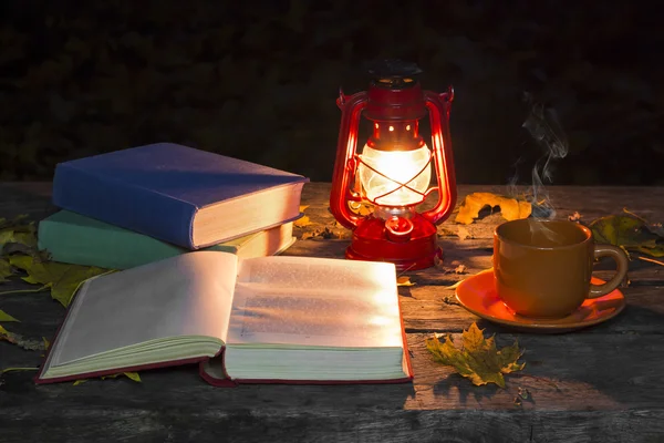 The open page of the book, lamp and a cup of hot coffee on the old wooden table in the dark forest. Fallen yellow maple leaves. Vintage and ancient concept. Autumn mood.