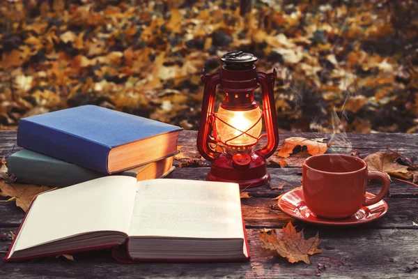 The book, lamp and a cup of hot coffee on the old wooden table in a forest. Fallen yellow maple leaves.