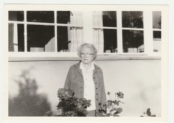 Vintage photo shows older woman in front of country house. Retro black & white  photography.