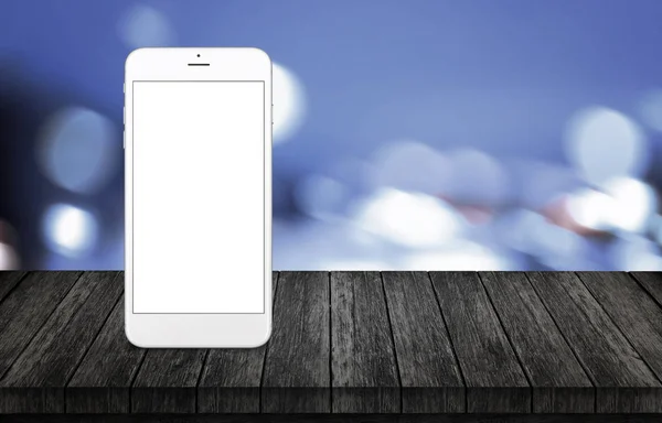 Smart phone on wooden desk. White phone with isolated, white, blank screen for mockup. Blurred city lights background.