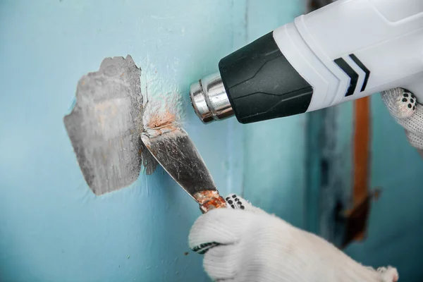 Master removes old blue paint from concrete wall with heat gun and scraper. Closeup