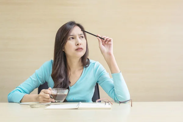 Closeup asian woman working with thinking face and a pencil and cup of coffee in her hand on blurred wooden desk and wall textured background in the meeting room , woman with thinking concept