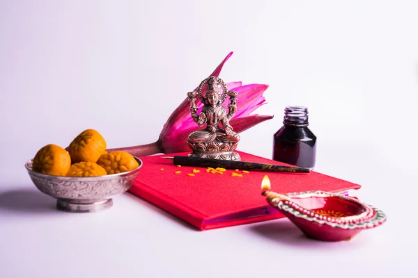 An auspicious Indian writing Shubha Labh means \'Goodness\' & \'Wealth\', over Red accounting note book / \'bahi khata\' with goddess Laxmi, diya, sweets and lotus and pen with ink on laxmi pujan, on diwali