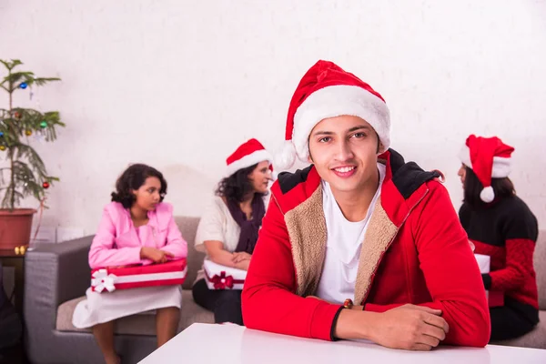 Indian young and smart man with red santa hat, sitting on table close to camera and family sitting on sofa in the background with christmas tree, Indians celebrating christmas, 25 december, merry christmas